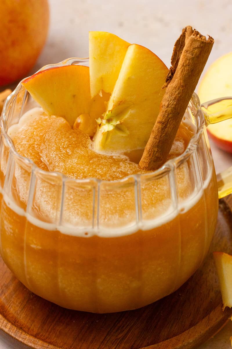 Close up of a frozen apple cider slushie in a glass mug with apple slices and cinnamon stick for garnishes.