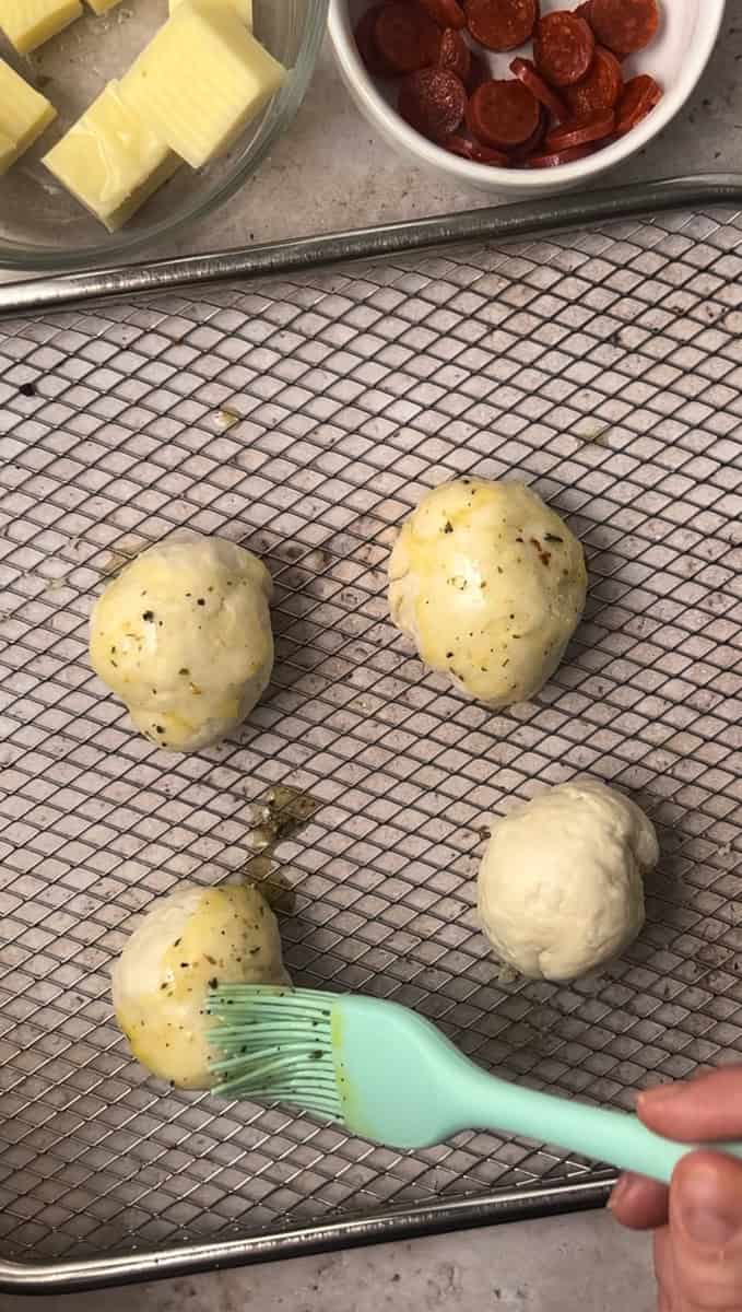 Four dough balls on a baking tray are being brushed with melted butter, next to bowls of sliced pepperoni and cubed cheese.