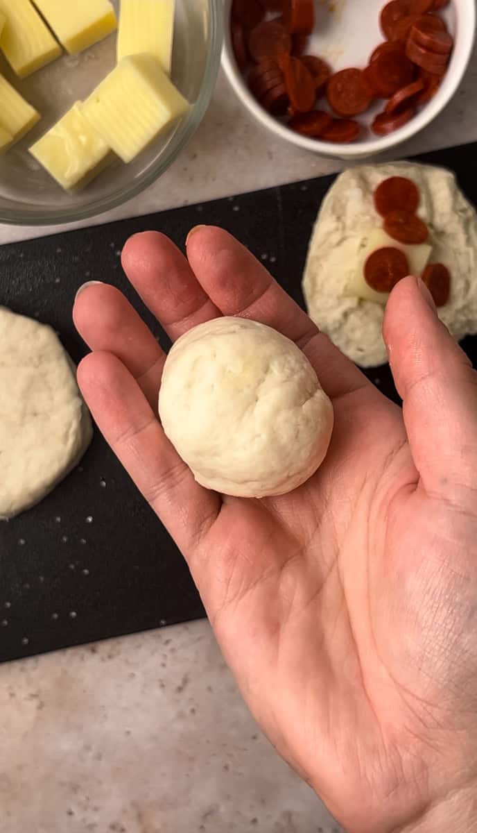 A hand holds a ball of dough. Nearby, there are bowls with cheese cubes and pepperoni slices. Another piece of dough with toppings is on a black cutting board.
