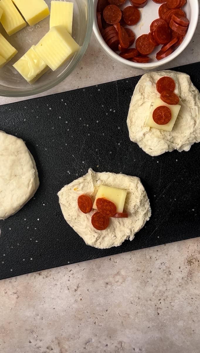 Dough pieces on a cutting board with slices of cheese and small pepperoni, next to a bowl containing more of the same ingredients.