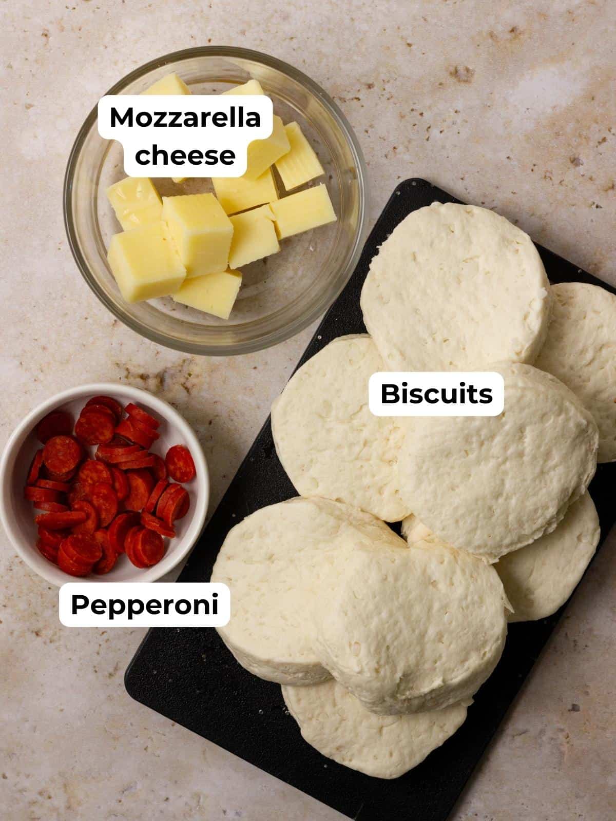 Ingredients for air fryer pizza bombs: cubed mozzarella cheese, slices of pepperoni, and uncooked biscuits on a black cutting board.