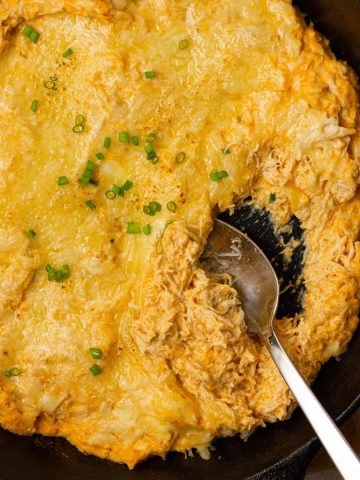 A skillet filled with creamy baked buffalo chicken dip with Greek yogurt, topped with melted cheese and chopped green onions. A spoon is scooping some out.