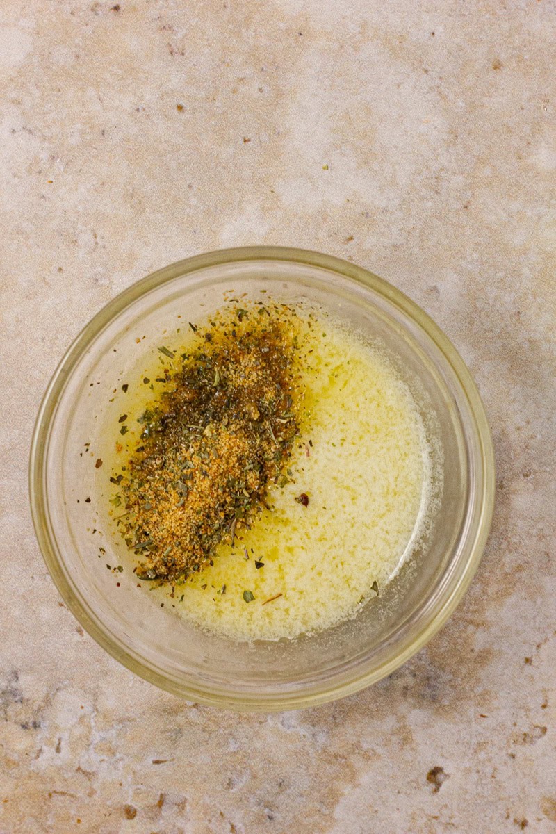 A glass bowl containing melted butter mixed with herbs and spices on a countertop.