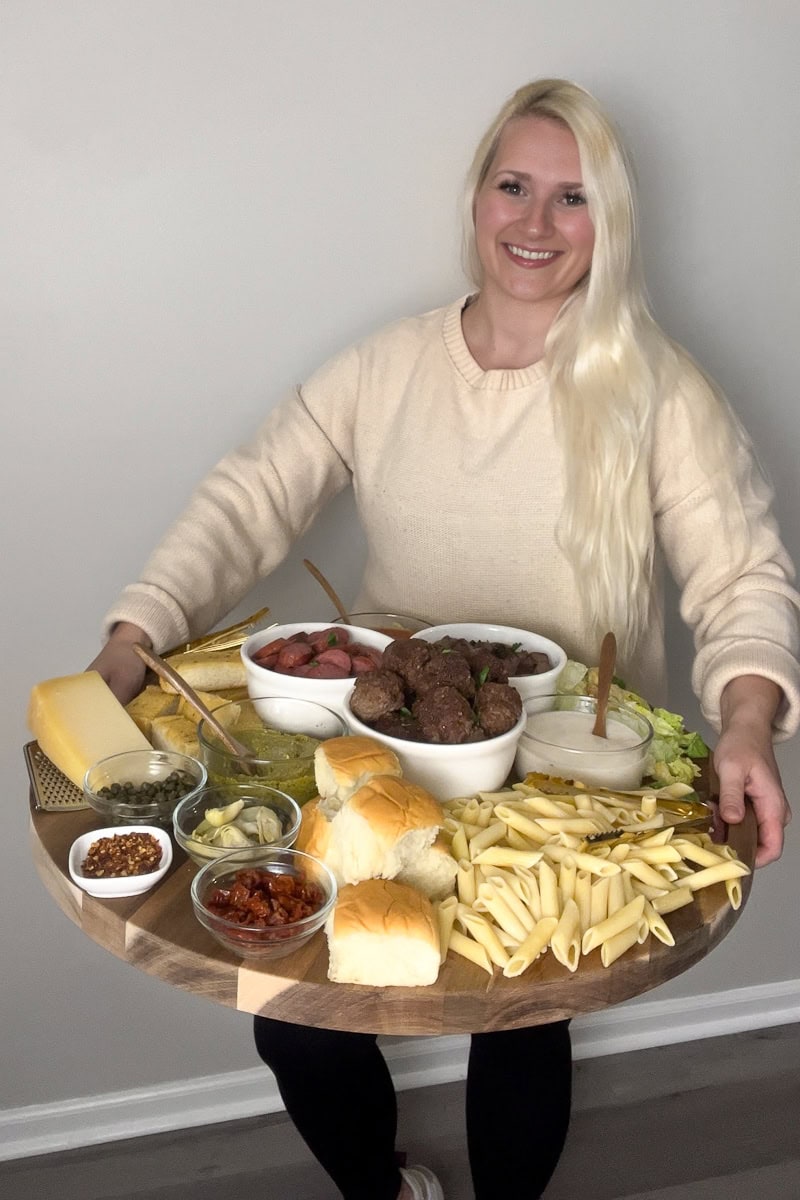 Blonde girl holding a large wooden platter with pasta, beef toppings, bread, and salad.