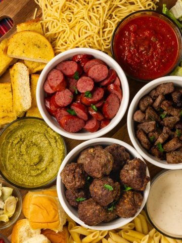 An epic pasta charcuterie board with spaghetti, penne, beef meatballs, sausage, steak tips, sauces, salad, and garlic bread.