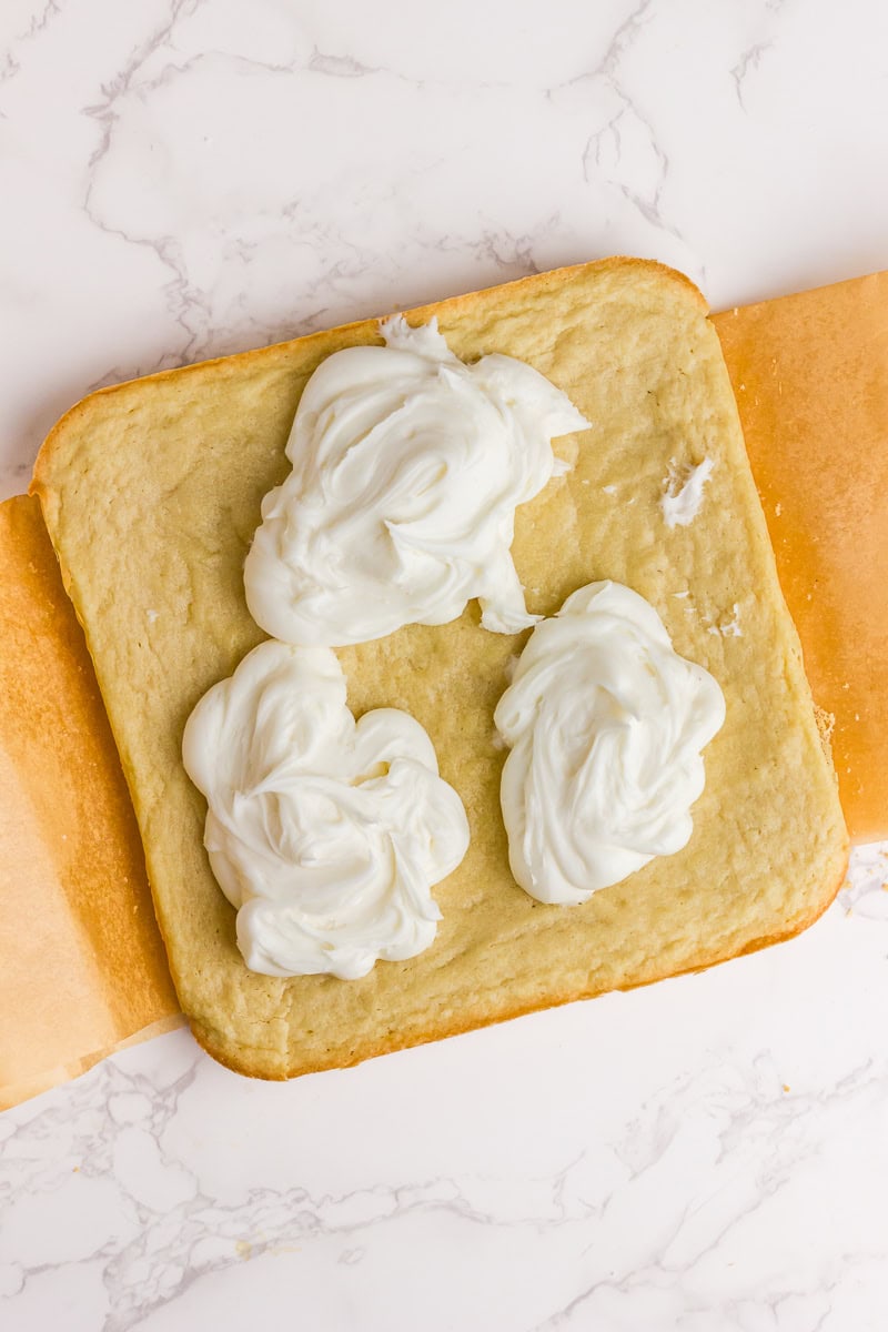 A baked cookie bar layer is topped with dollops of white frosting before being spread.
