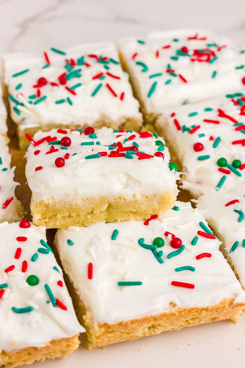 A close-up of frosted sugar cookie bars shows a soft interior and colorful holiday sprinkles on top.