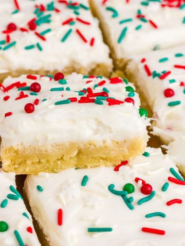 Frosted Christmas sugar cookie bars are stacked and topped with red, green, and white sprinkles.