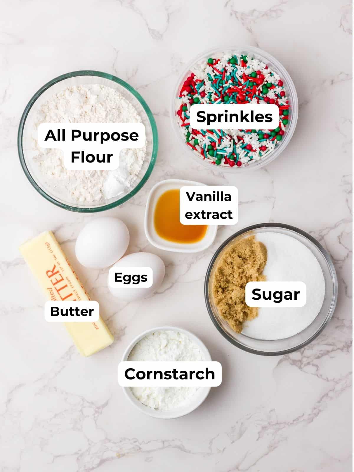 Ingredients for Christmas sugar cookie bars are arranged on a marble surface, including flour, sugars, eggs, butter, sprinkles, extracts, and cornstarch.