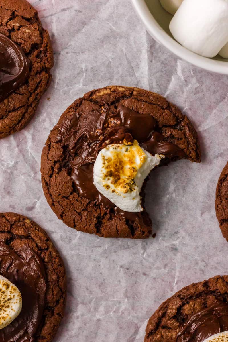 A partially eaten chocolate cookie with a toasted marshmallow on top, surrounded by other cookies.