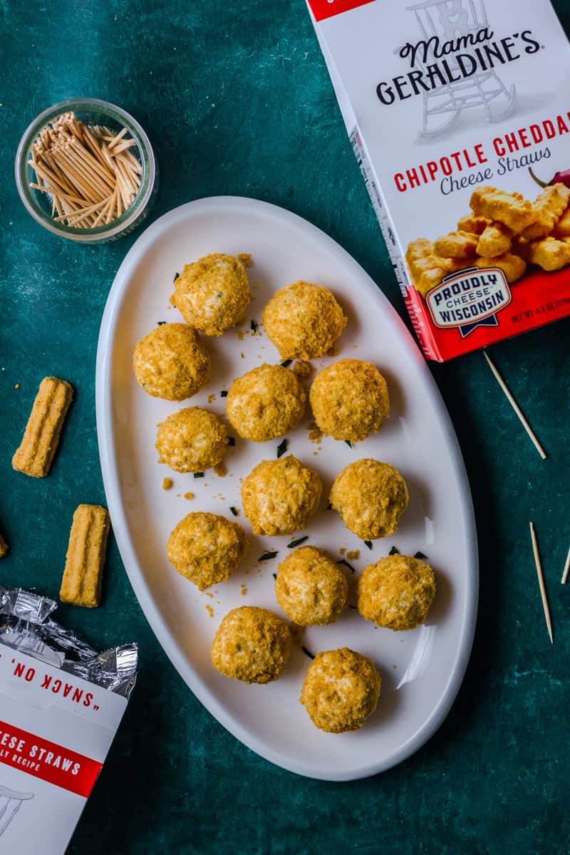 A white plate filled with mini cheese balls coated in crushed Mama Geraldine's Chipotle Cheddar Cheese Straws, placed on a green background.