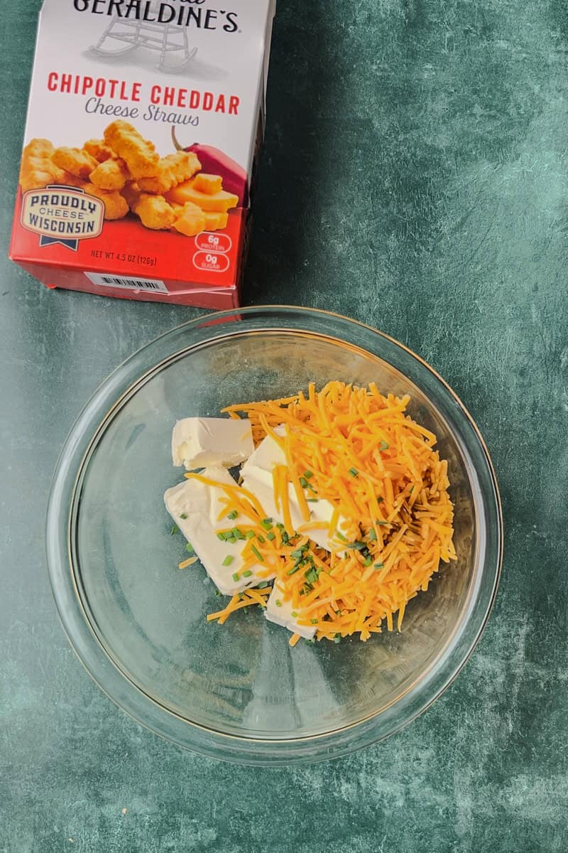 A glass bowl filled with cream cheese, shredded cheddar, and chives, next to a box of Mama Geraldine's Chipotle Cheddar Cheese Straws.