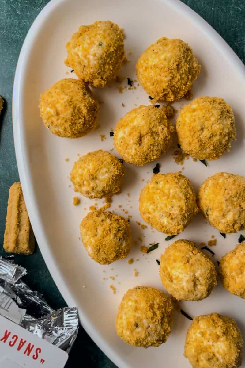 A close-up of mini cheese balls coated in crushed cheese straws, arranged on a white plate with crumbs and garnishes.