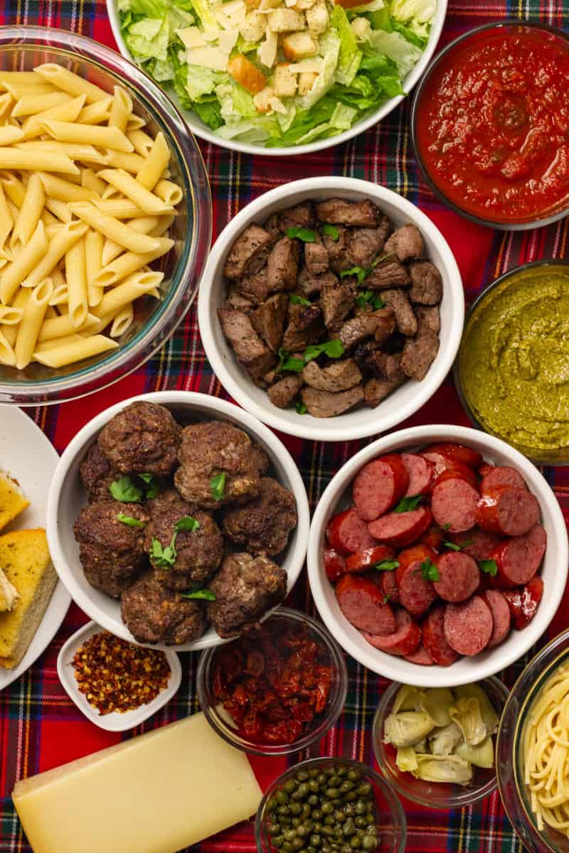 Ingredients to build a pasta board with various beef, pasta, and sauce options on a red and green plaid table cloth.