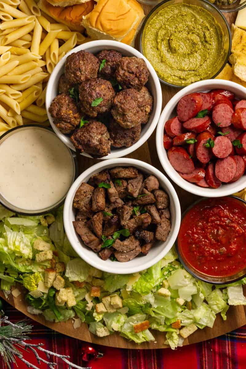 A festive pasta charcuterie board showcasing beef meatballs, sausage, steak tips, pasta, sauces, and Caesar salad.