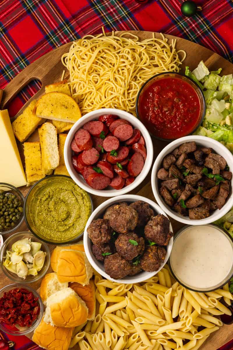Festive pasta charcuterie board with spaghetti, penne, beef meatballs, sausage, steak tips, sauces, garlic toast, and toppings, set on a wooden board with a holiday plaid backdrop.