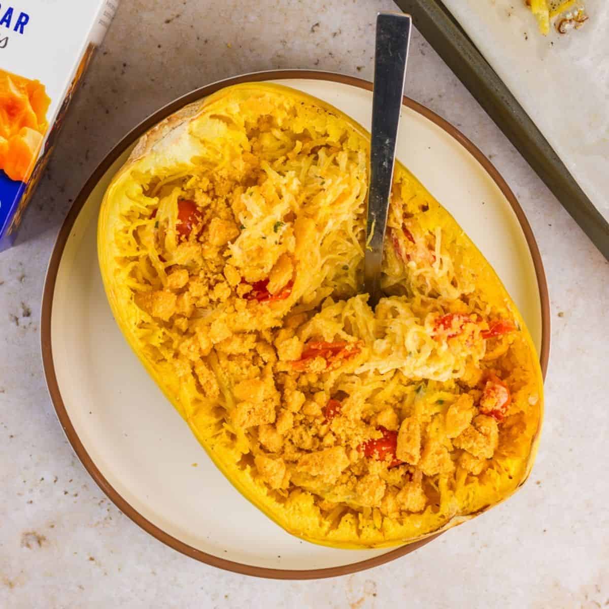 Halved cooked spaghetti squash filled with shredded squash, cheese straw crumbs, and cherry tomatoes, with a fork inserted, placed on a baking sheet.