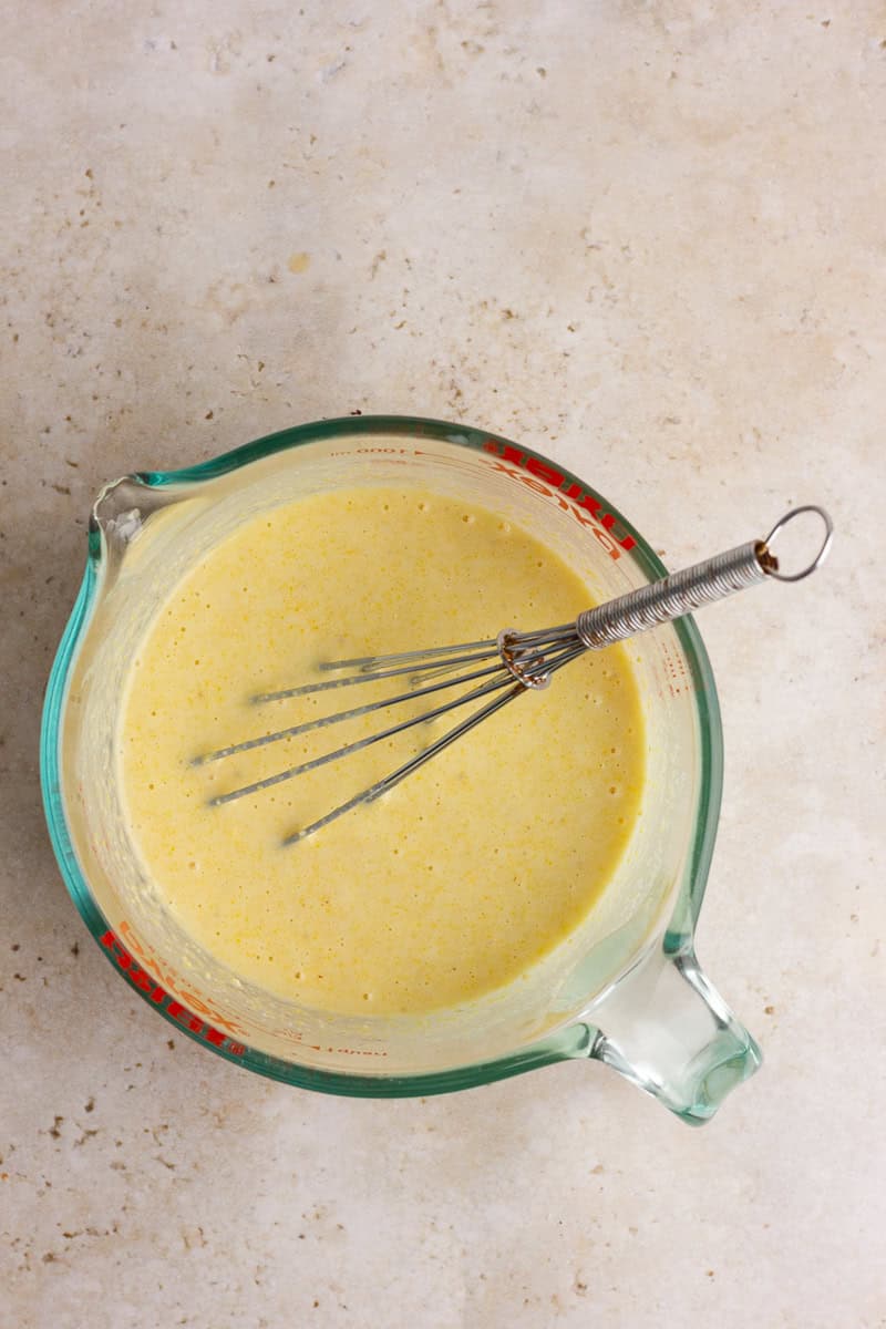 A glass measuring cup filled with cornbread batter and a whisk on a neutral countertop.