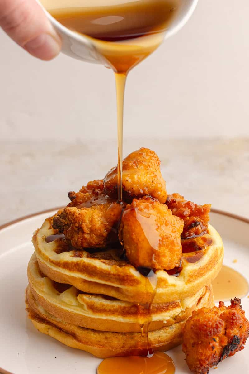 A stack of cornbread waffles topped with crispy chicken nuggets, drizzled with syrup.