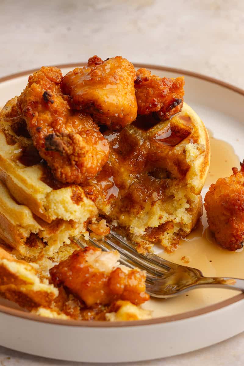 A close-up of cornbread waffles and chicken, cut open with a fork, showing the fluffy texture and syrup.