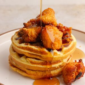 A featured shot of stacked cornbread waffles with crispy chicken and syrup pouring over the top.
