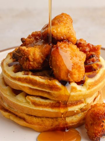A featured shot of stacked cornbread waffles with crispy chicken and syrup pouring over the top.