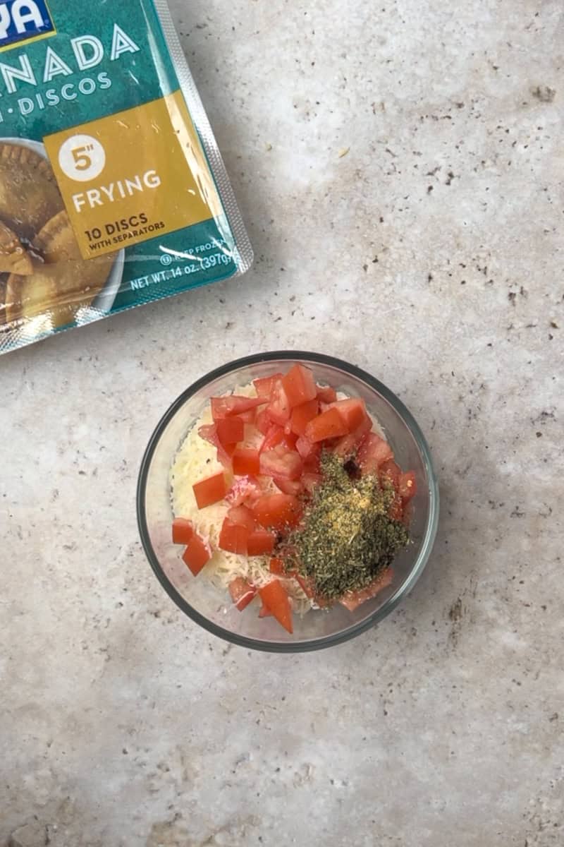 A bowl of diced tomatoes, shredded mozzarella, and Italian seasoning next to a package of empanada discs.