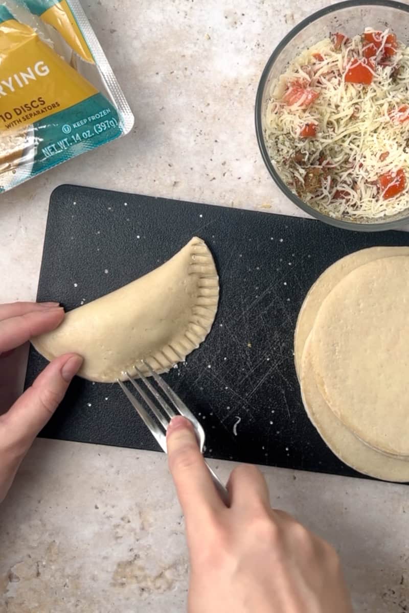 A hand using a fork to crimp the edges of a filled empanada on a black cutting board.