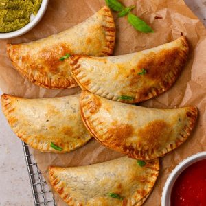 Crispy, golden-brown empanadas caprese on parchment paper, served with fresh basil, pesto, and marinara dipping sauce.