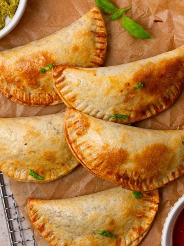 Crispy, golden-brown empanadas caprese on parchment paper, served with fresh basil, pesto, and marinara dipping sauce.