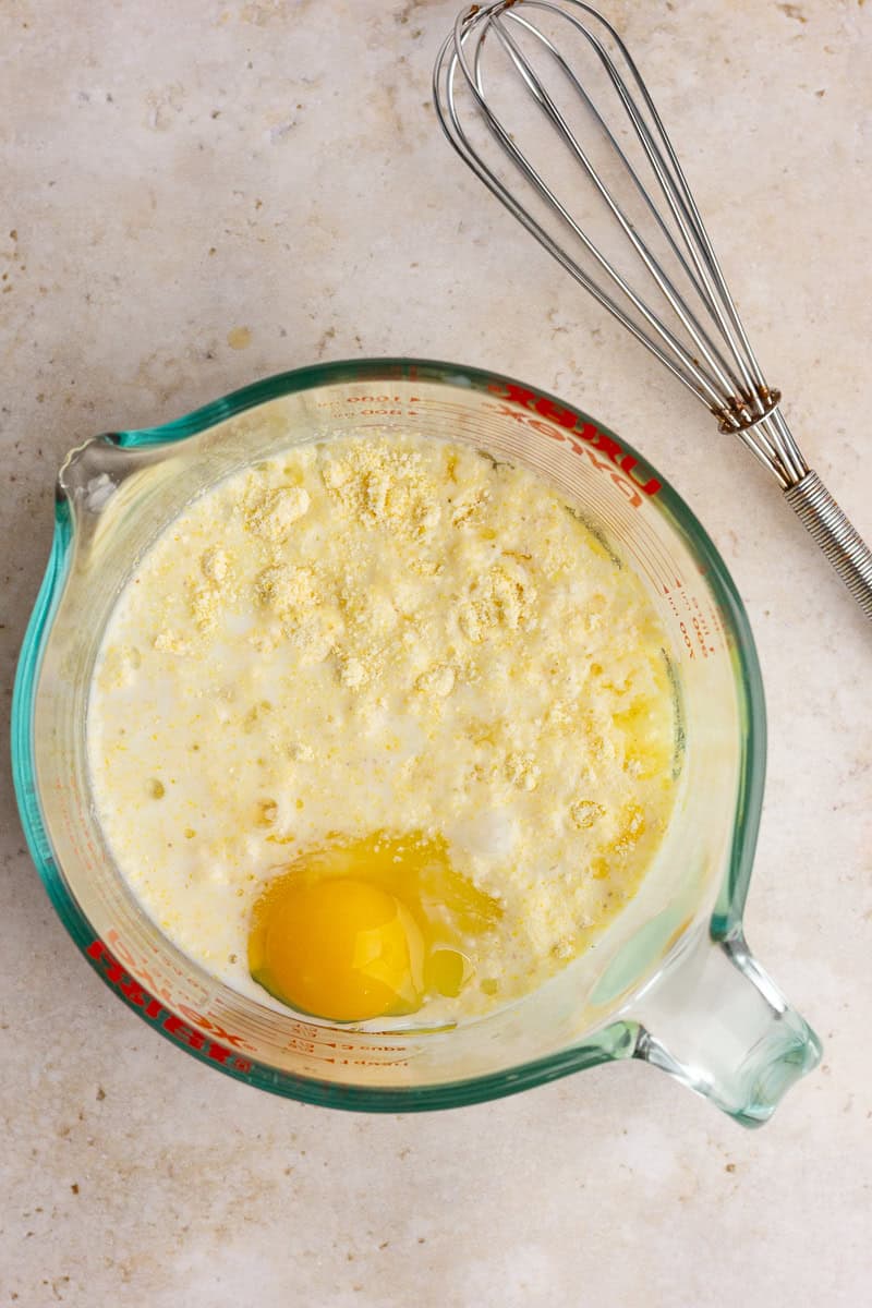 A glass measuring cup with Jiffy cornbread mix, milk, an egg, sugar, and oil, ready to be mixed.