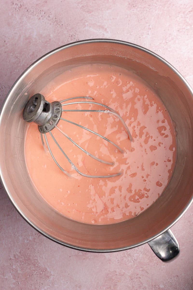 A mixing bowl with a pink batter and a whisk attachment inside.