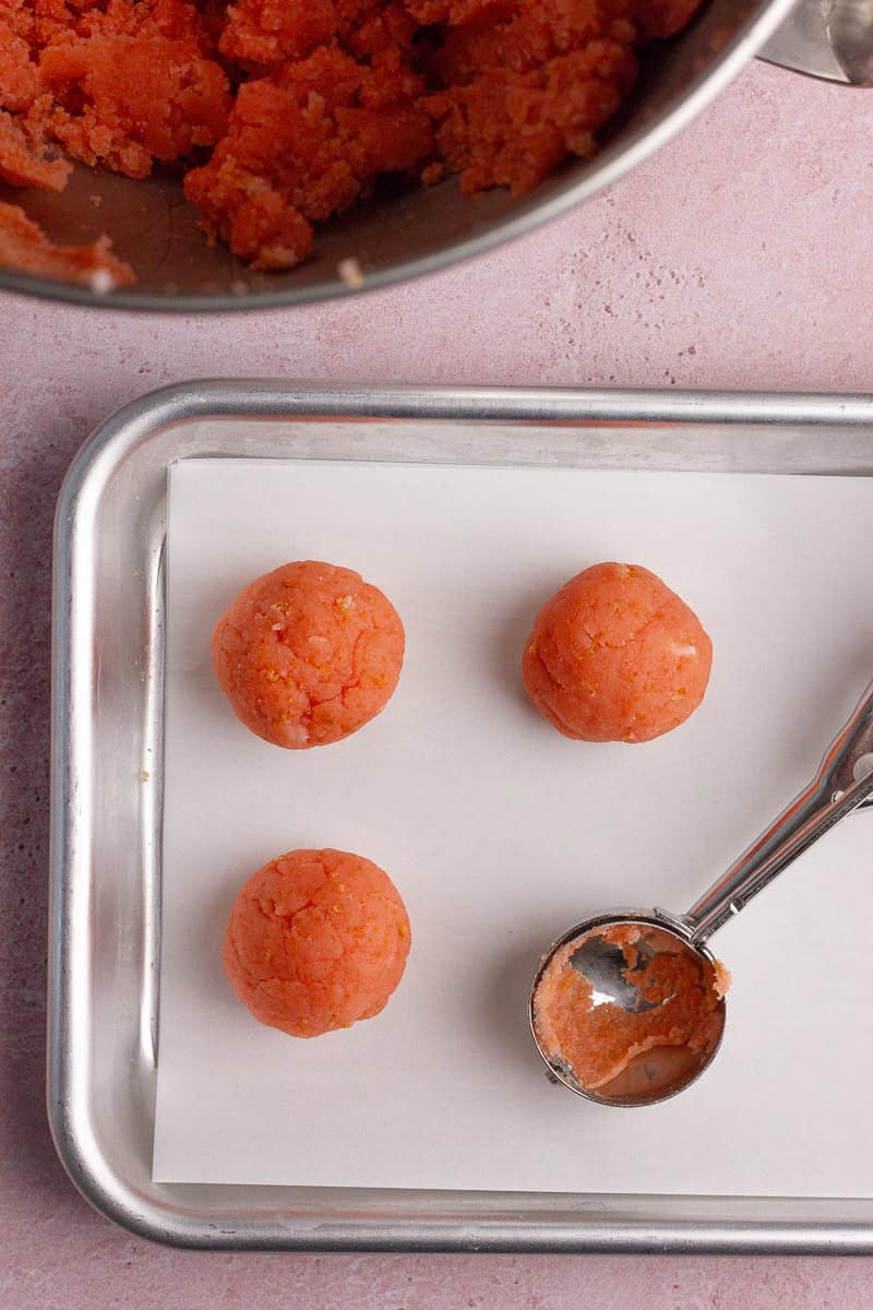 A baking sheet lined with parchment paper, holding three rolled strawberry cake balls and a cookie scoop.