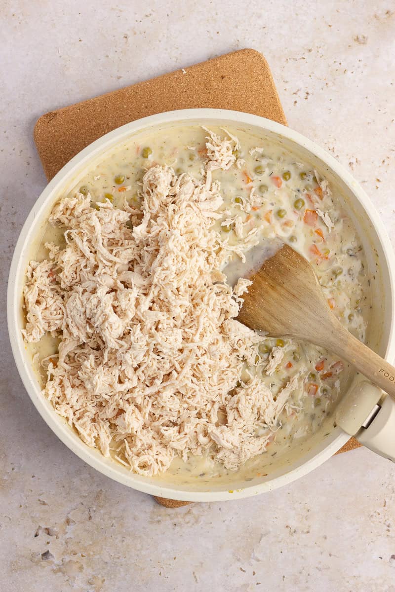 Shredded chicken being mixed into a creamy pot pie filling with peas and carrots.