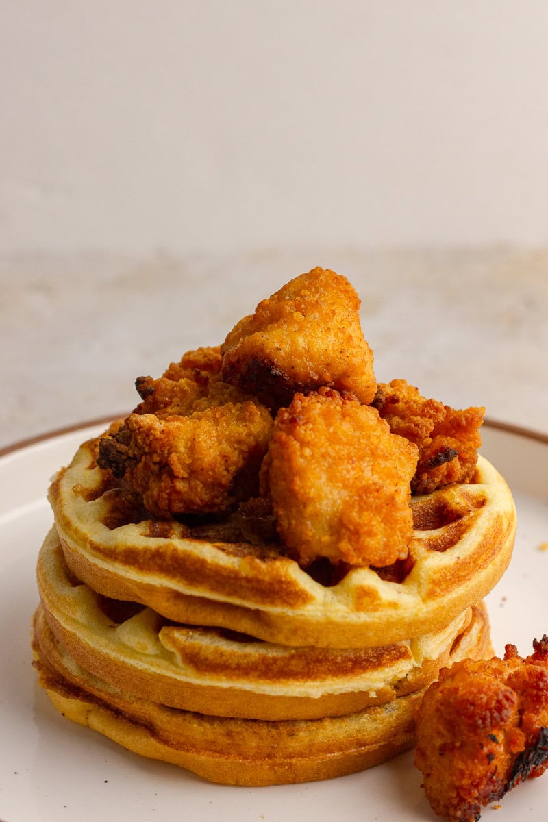 Stack of cornbread waffles topped with crispy chicken nuggets on a white plate.