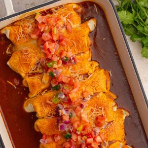A close-up of baked enchiladas covered in red sauce, melted cheese, and fresh pico de gallo, with cilantro on the side.