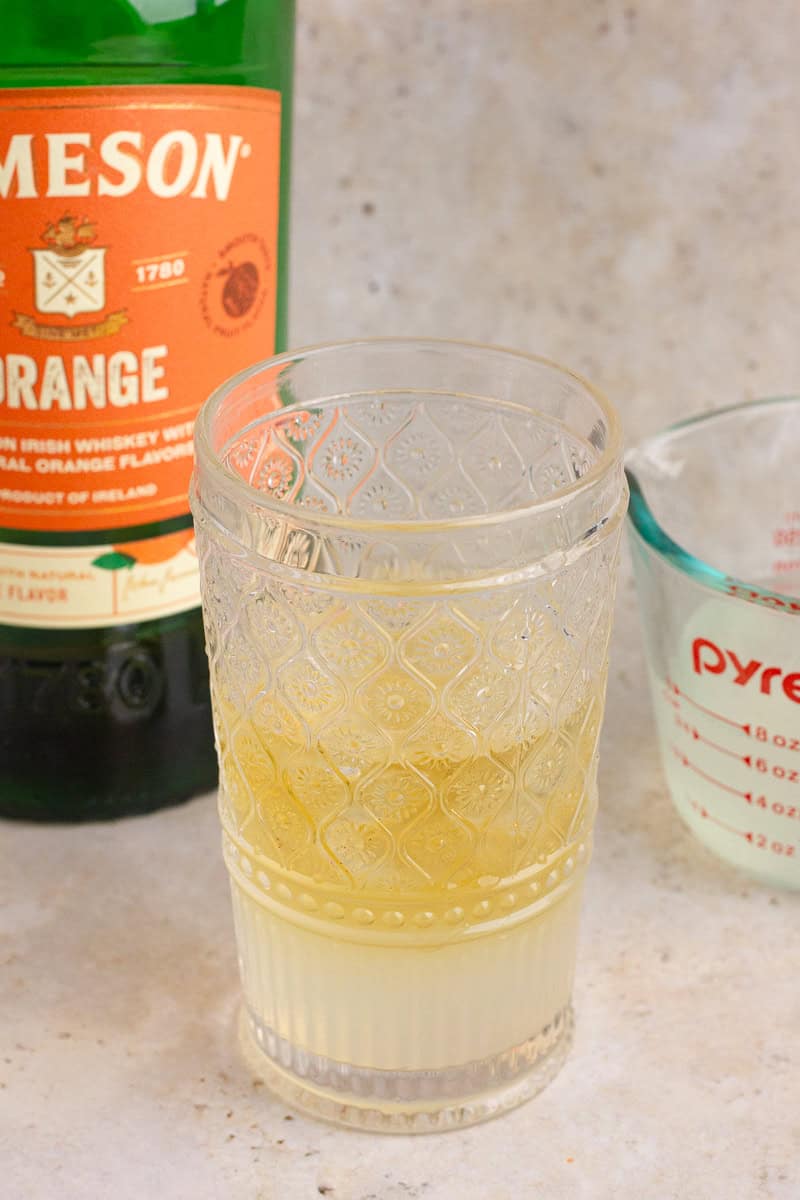 A cocktail shaker with Jameson Orange and lemonade mixture, with a Jameson Orange bottle and a measuring cup in the background.