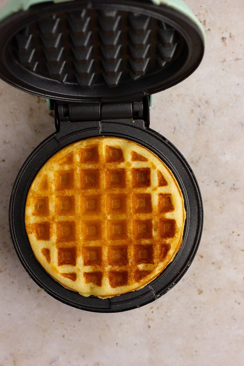 A golden-brown cornbread waffle cooking in a mini waffle maker.