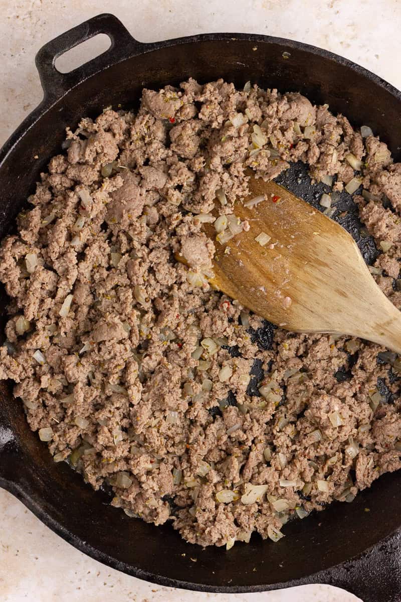 A close-up of turkey sausage being cooked in a cast-iron skillet with diced onions and Italian seasoning.