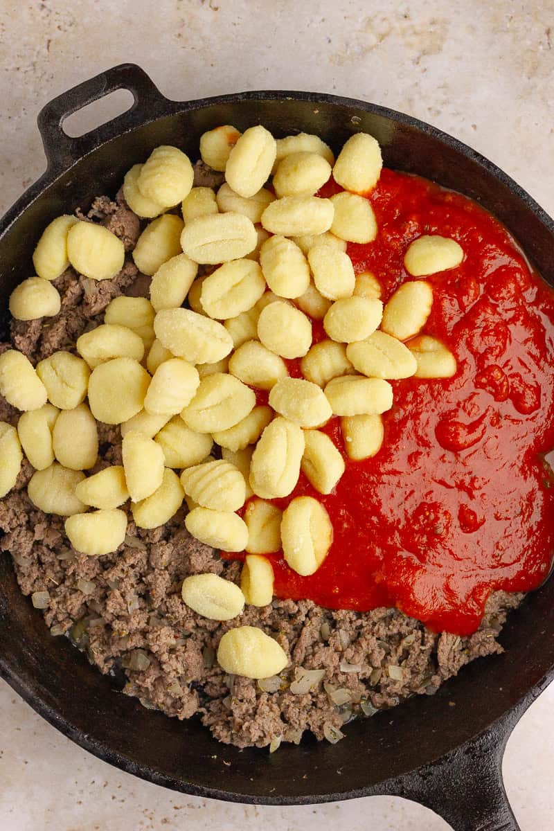 A cast-iron skillet filled with cooked sausage, uncooked gnocchi, and marinara sauce, ready to be mixed.