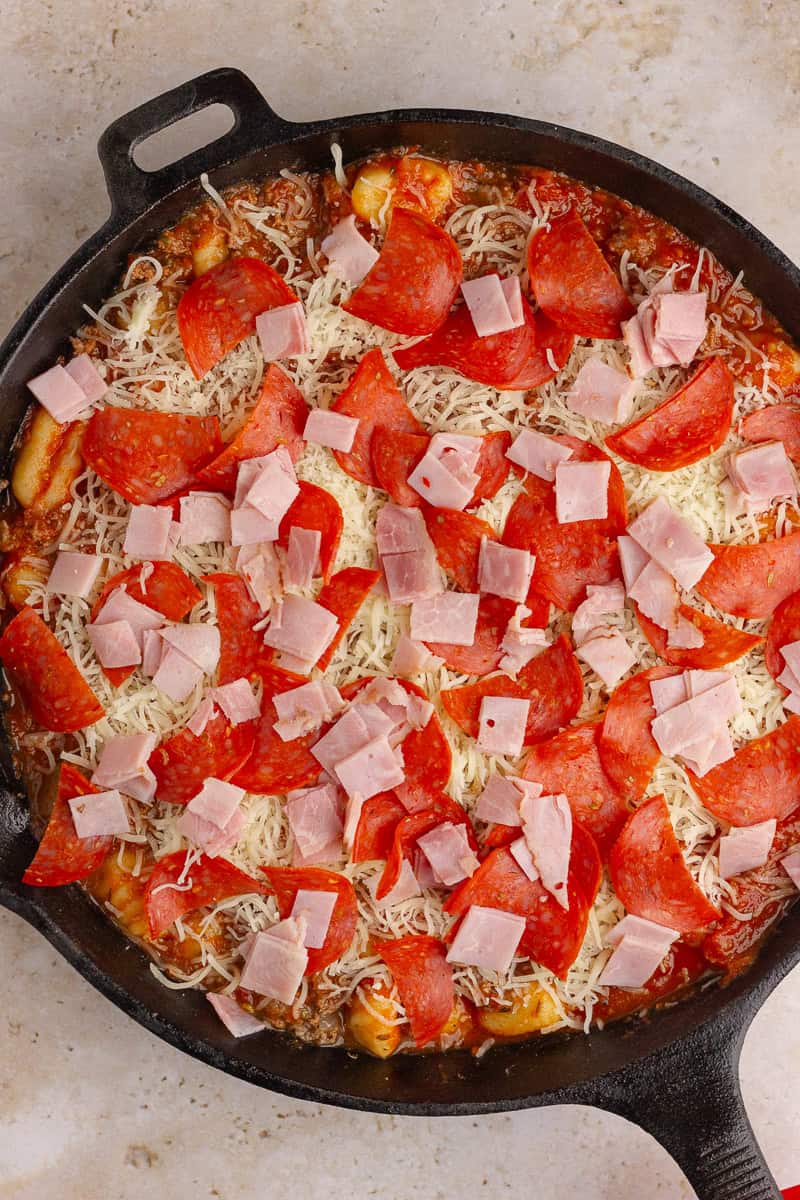 The assembled Pizza Gnocchi dish before baking, topped with shredded mozzarella, pepperoni slices, and diced ham.