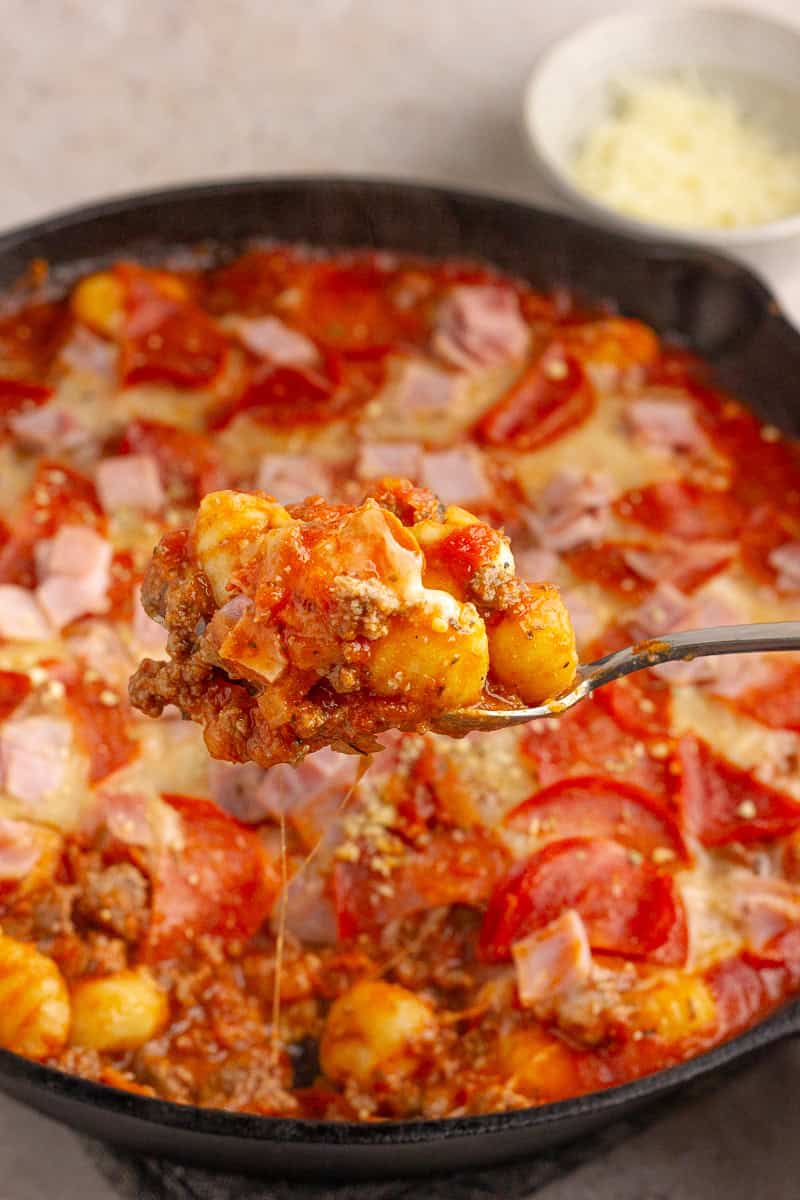 A close-up of a spoonful of cheesy, saucy Pizza Gnocchi, showcasing tender gnocchi, savory sausage, and gooey melted cheese with crispy pepperoni in the background.