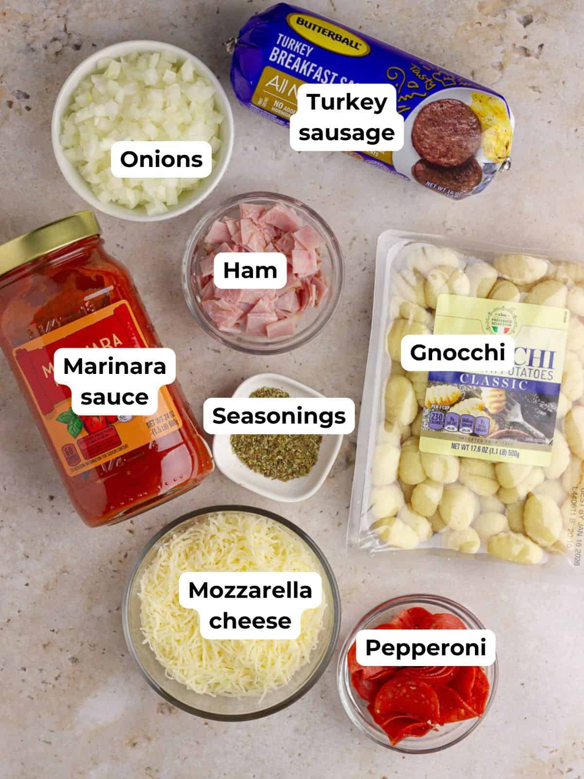 A labeled flat-lay image of all the ingredients for Pizza Gnocchi, including mozzarella cheese, marinara sauce, turkey sausage, seasonings, gnocchi, onions, pepperoni, and ham.