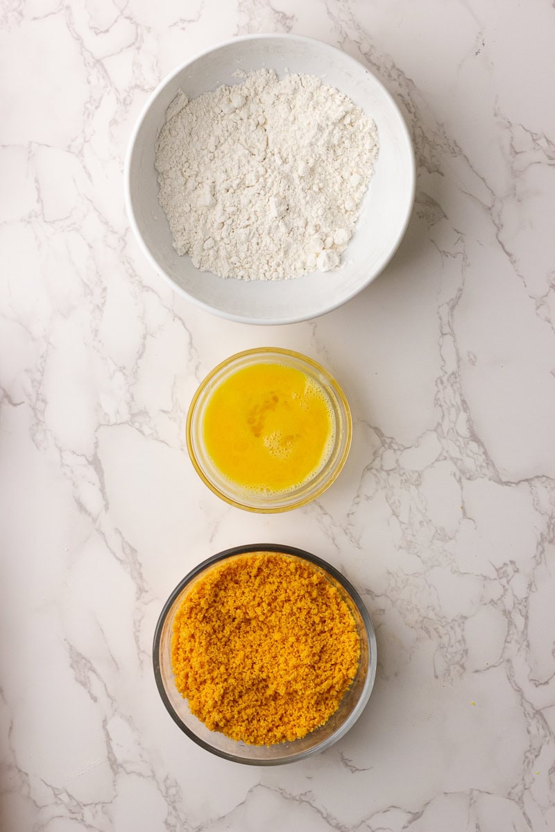 Three bowls showing flour, beaten eggs, and crushed cheese straws for coating deviled eggs.