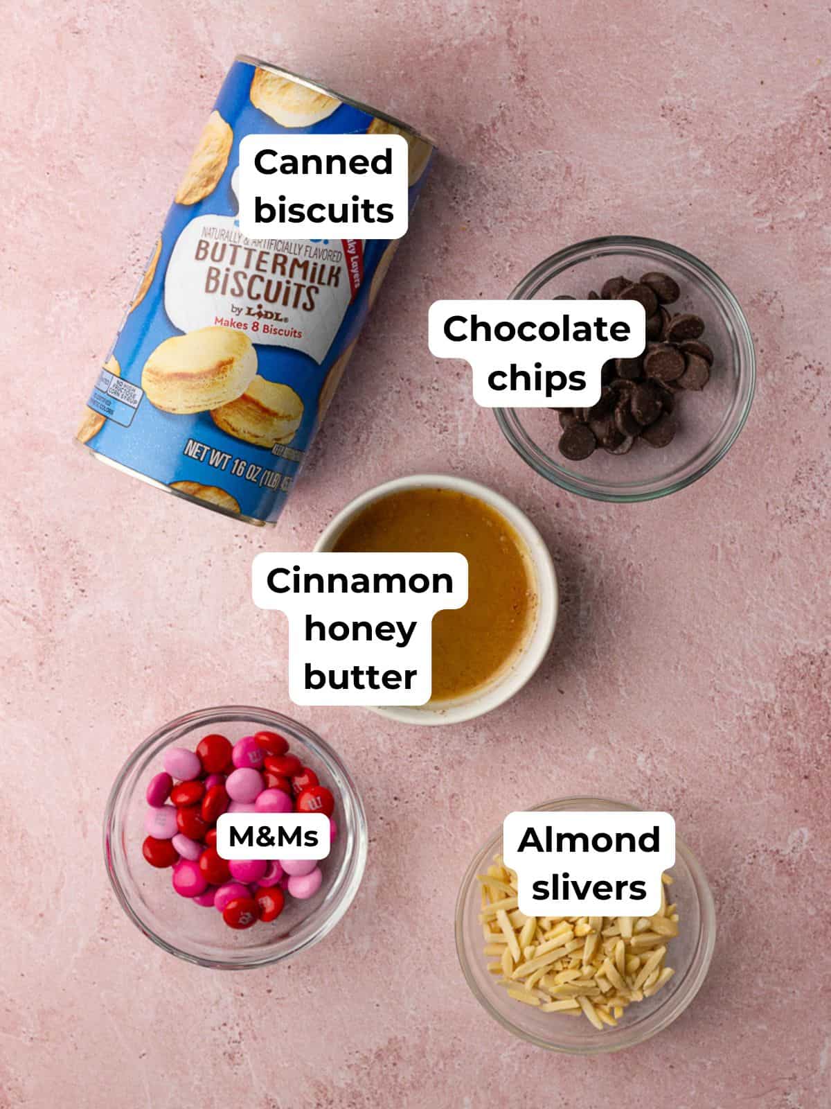 Overhead of ingredients for Easter Bunny Biscuits including canned biscuits, M&Ms, chocolate chips, almond slivers, and cinnamon honey butter.