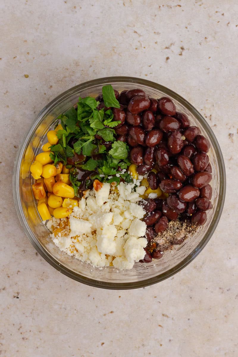 Ingredients layered in a clear bowl before mixing: black beans, corn, feta, cilantro, and seasoning.