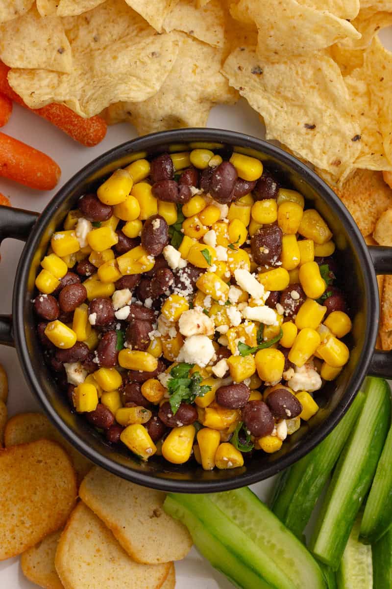 Black bean corn feta dip served in a black bowl surrounded by chips, carrots, and cucumbers.