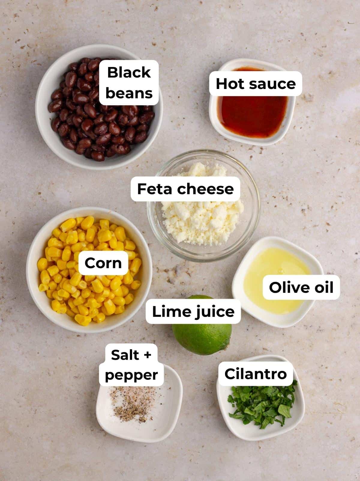 Labeled ingredients for the dip laid out on a counter, including beans, corn, feta, lime, and seasonings.