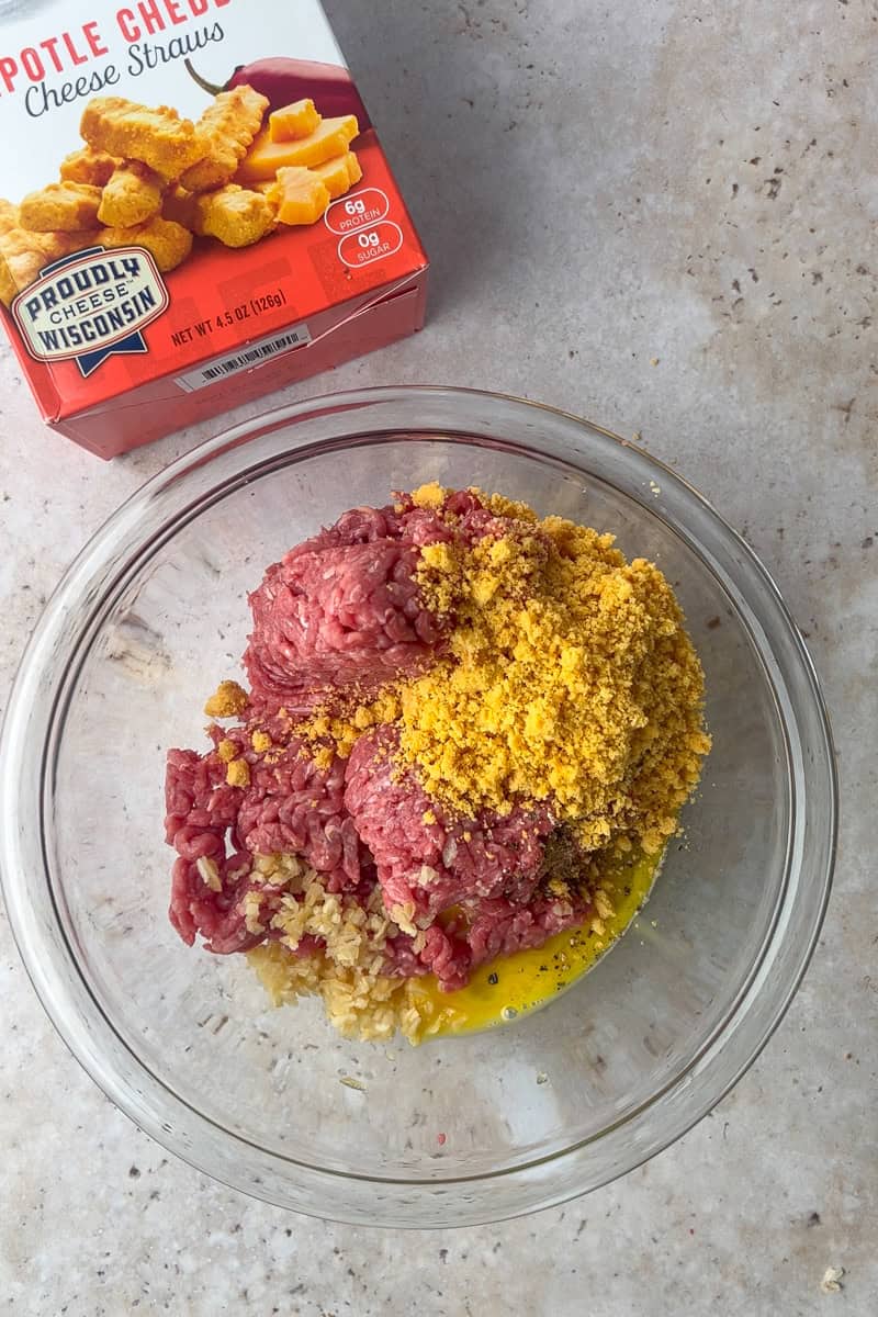 Ground beef, garlic, egg, onion, and crushed chipotle cheese straws in a clear mixing bowl, ready to be combined for burgers.