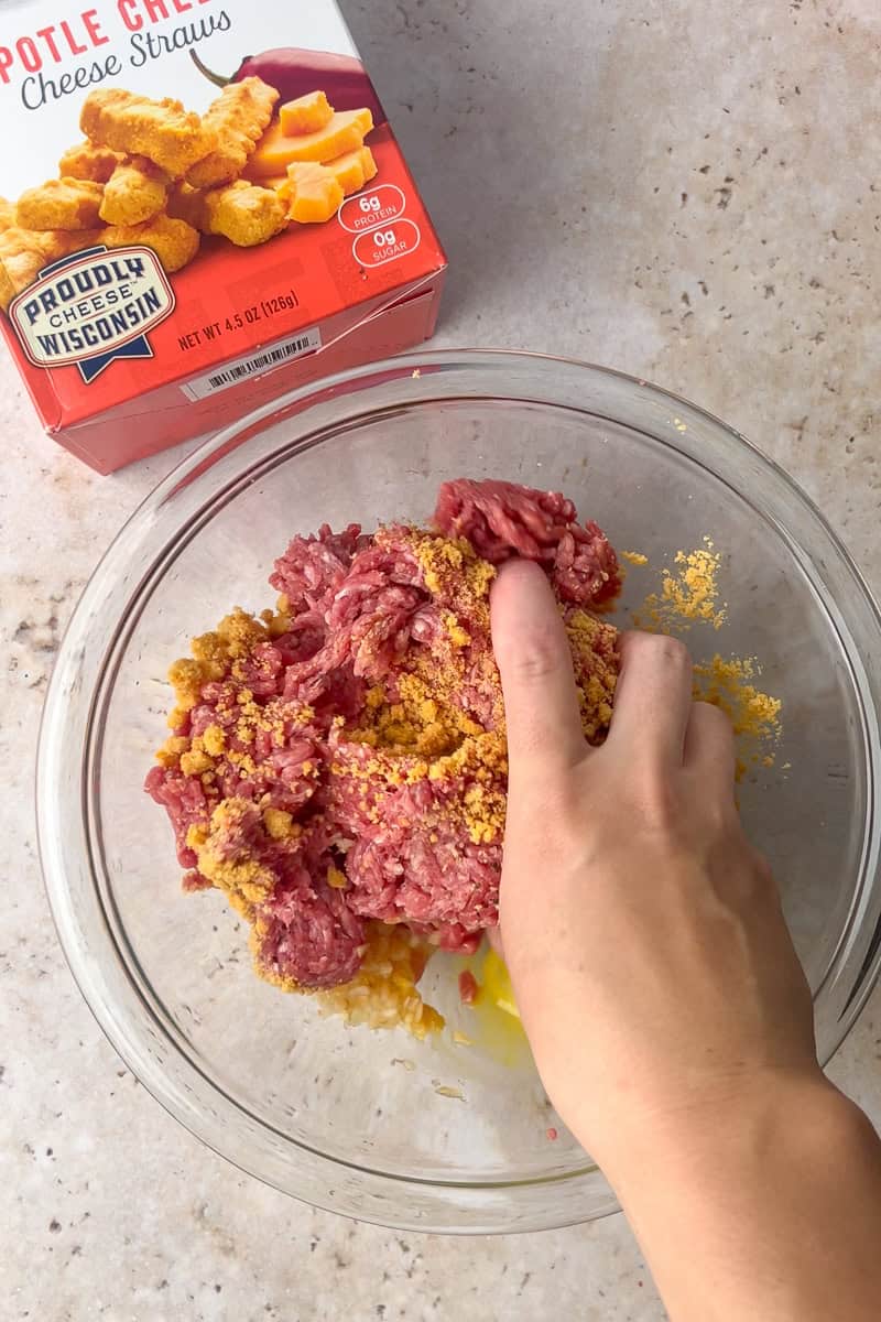 A hand begins mixing together the burger ingredients in a glass bowl with Mama Geraldine's cheese straws in the background.